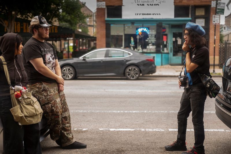 Uriel Reyes, photographs two young Pilsen residents on W. 18th St. in Pilsen, Chicago during the 2026 National Geographic Photo Camp. Uriel student on the right, wearing a blue beret and glasses, holds a camera and appears to be observing or discussing. Another student, wearing a baseball cap and camouflage pants, leans against a vehicle, while a third student, wearing a dark hoodie and carrying a canvas bag, stands nearby. In the background, a dark sedan is parked in front of a storefront with a 'Mermaid Spa Nail Salon' sign. This photograph documents a moment during the National Geographic Photo Camp 2026, held from August 9-14, which provided photography training to Yollocalli Arts Reach students.