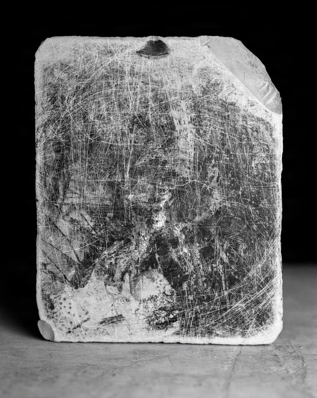 A close-up, black and white, vertical studio shot captures the heavily scratched and damaged surface of what appears to be a Plattenkalk specimen. The rectangular object, possibly a fossil slab, exhibits numerous fine, intersecting white lines indicative of deep scratches across its dark, mottled surface. A small, dark, triangular indentation is visible near the top edge, and the top right corner shows a chipped or broken area. The bottom left corner is rounded, and the object rests on a lighter, textured surface, possibly a table or floor. The background is solid black.