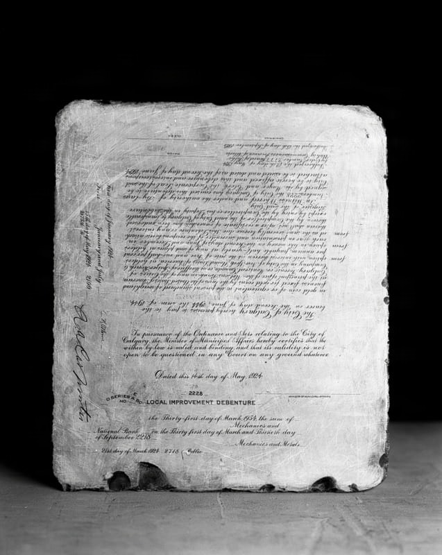 A close-up, high-angle view of a Local Improvement Debenture document from the City of Calgary, dated May 14, 1924. The document, likely a lithography stone, displays text detailing financial obligations, including dates like 'First day of January 1924' and 'Second day of June 1944,' and references to 'The City of Calgary' and 'Minister of Municipal Affairs.' Signatures or spaces for signatures for 'Mayor,' 'Clerk,' and 'Chairman' are visible. The document shows signs of age and wear, with visible scratches and chipped edges, particularly at the bottom. This image is part of David Christensen's 'Plattenkalk' series, created in 2026.