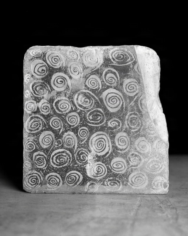 A close-up, black and white studio shot captures a rectangular slab of Plattenkalk, a fine-grained limestone, standing upright. The surface of the stone is densely covered with numerous spiral patterns of varying sizes, appearing as lighter forms against a darker background. The texture of the stone is visible, with some rough edges and a slightly irregular top surface. The slab rests on a flat, light-colored surface, with a dark background behind it.