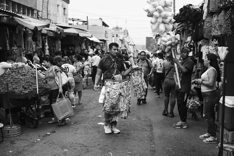 Bustling outdoor market scene with vendors selling clothes and food, crowded with people shopping and interacting on a street lined with stalls.