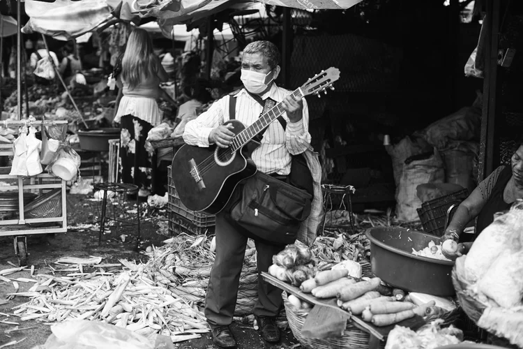 A man wearing a mask playing a guitar at an outdoor market, with various vegetables displayed nearby and people in the background.