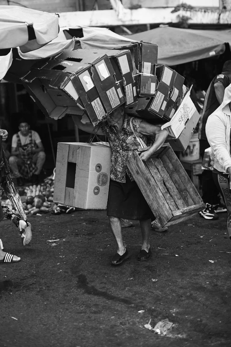 Person carrying a large stack of cardboard boxes and a wooden crate in a busy market setting.