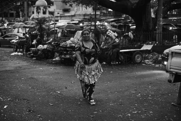 A black and white street photograph showing a woman in a dress walking while holding a bouquet of flowers. She is in an outdoor market setting with various stalls and vehicles in the background. The mood is casual and lively with market activity.