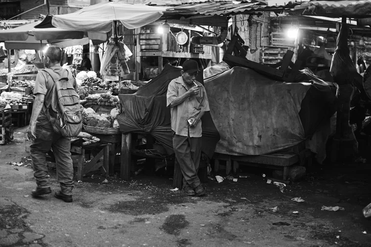 Black and white photo of a bustling outdoor market with a man smoking and another walking by, surrounded by various covered stalls and produce.
