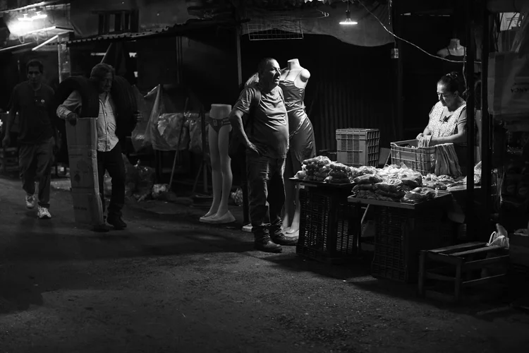 Outdoor night market scene with a man carrying tires, another man near mannequins, and a woman arranging items on a stall.