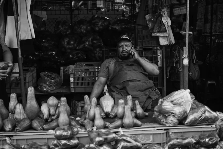 Black and white photo of a vegetable vendor talking on a phone, surrounded by crates and baskets of produce, likely squashes or gourds, at a market stall.