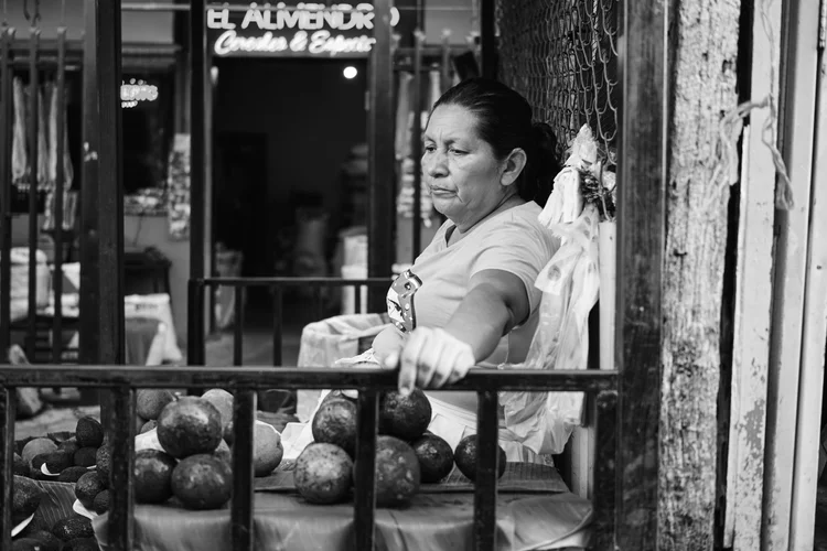 Black and white photo of a woman selling avocados at an outdoor market.