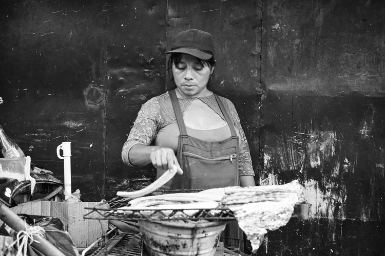 Black and white photo of a person wearing an apron and hat, grilling food on a street stand.