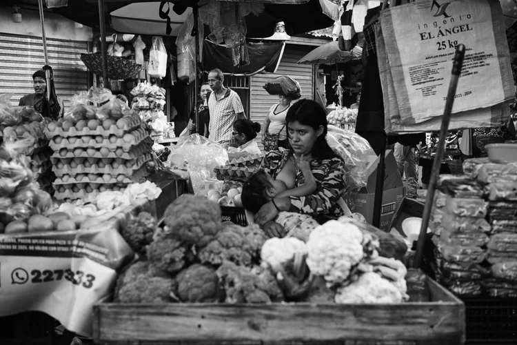 Black and white photo of a busy outdoor market with people, including a woman breastfeeding a child, surrounded by vegetables and stacked egg cartons.