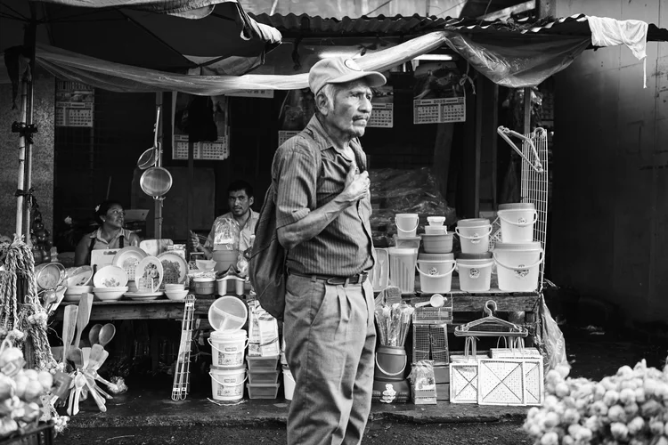 Black and white image of an elderly man standing in front of a market stall displaying kitchenware and plastic containers.