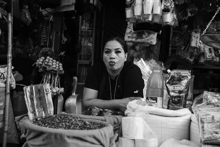 Black and white photo of a vendor at a market stall. The vendor is sitting behind bags of goods, including beans and other food items. Various packaged products and hanging goods are visible in the background.