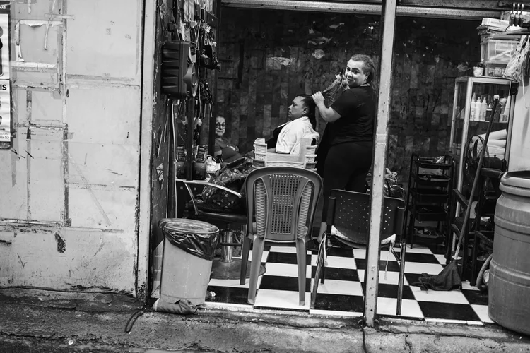 Black and white photo of a small barbershop with checkerboard flooring, a woman styling a customer's hair, another person sitting, and various hair products and chairs around.