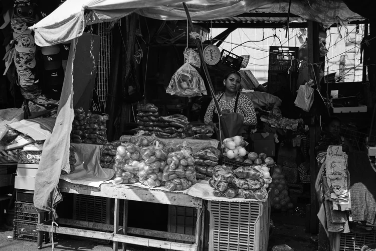 Black and white photo of a market stall with fruits and vegetables, bags of produce, a person wearing an apron standing behind a scale, and various items on shelves under a tarpaulin.