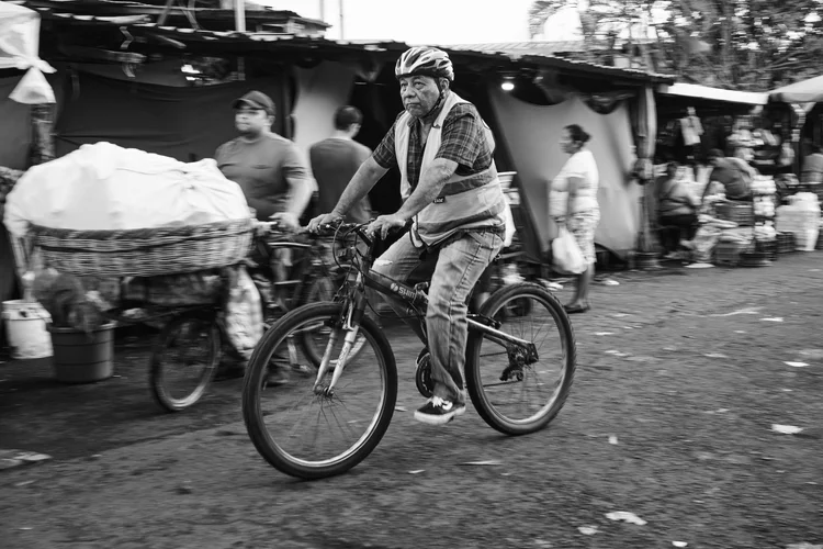 Black and white street scene with a man on a bicycle wearing a helmet and a reflective vest; market stalls and people in the background.