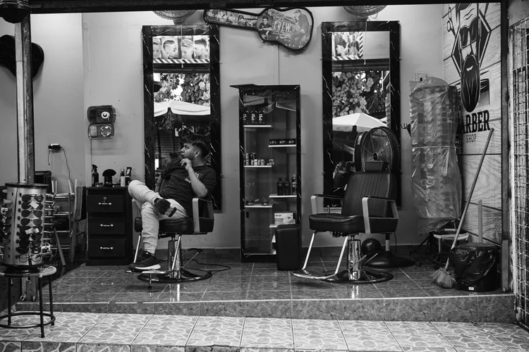 Black and white photo of a barbershop interior with a man sitting on a barber chair, mirrors, hair products, and a guitar-shaped sign on the wall.