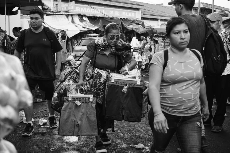 Black and white market scene with a woman selling clothing items like shorts; other people walking in various directions.