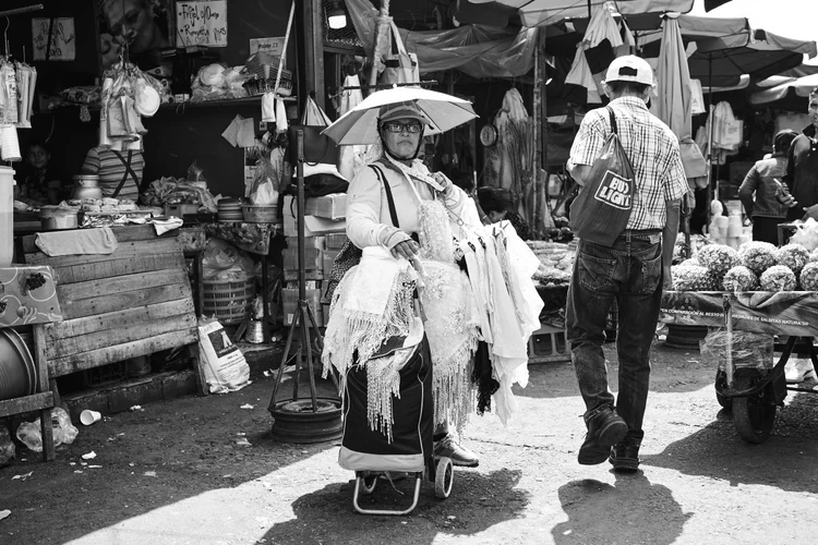 Black and white photo of a bustling market with a woman holding an umbrella and selling lace garments, standing beside a man carrying a Bud Light bag.