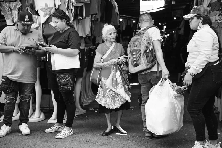 A black and white photo of five people in a busy market, with clothing displayed in the background. Two individuals are holding phones, while an elderly woman carrying a bag and cloth is flanked by people with a backpack and a large shopping bag.