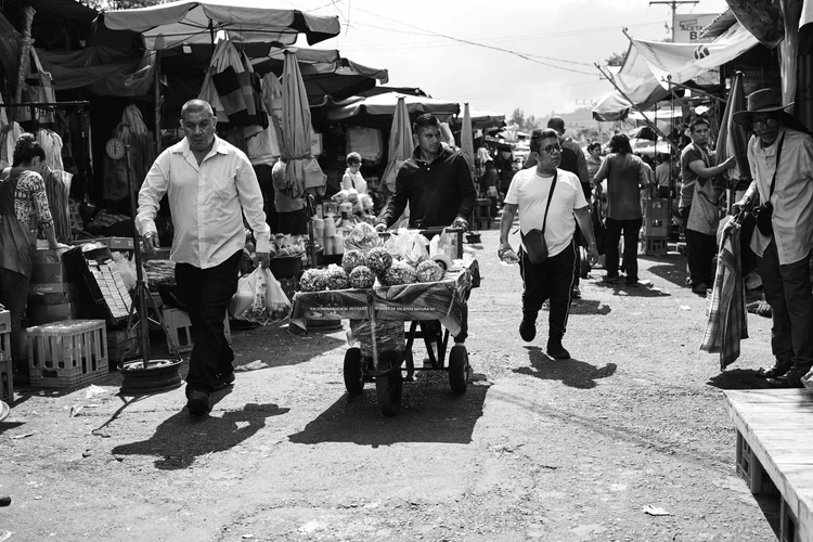 People walking through a busy outdoor market, with a vendor pushing a cart filled with various goods, surrounded by stalls and shoppers.