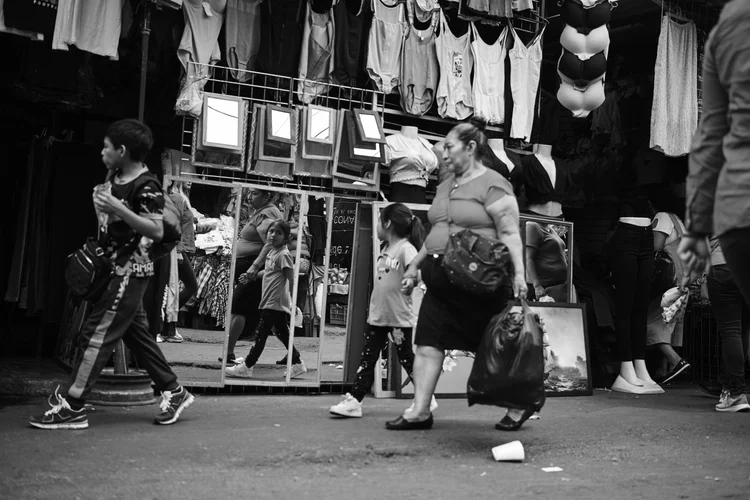 Street market scene with people passing by mirrors and hanging clothes, including t-shirts, camisoles, and bras.