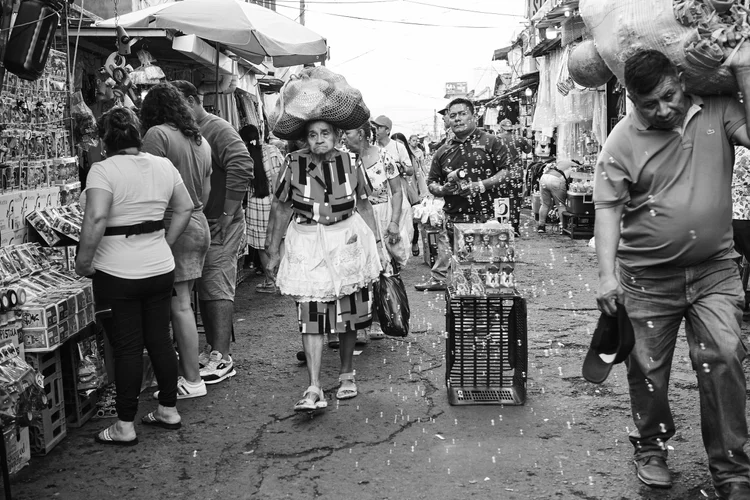 Black and white photo of a busy outdoor market street with people shopping and carrying goods. A woman is wearing a dress and apron, carrying a bag, and bubbles are visible in the air.