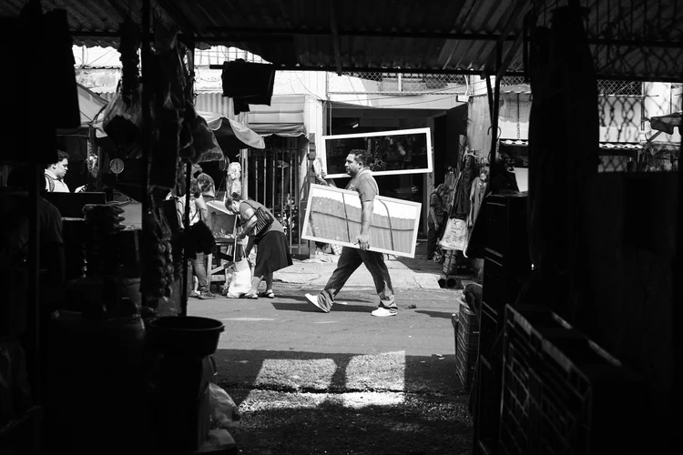 Black and white photo of a man carrying large frames through a busy outdoor market, with people shopping and stalls in the background.