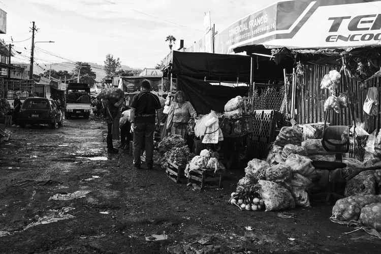 Black and white photo of a street market with people, produce stalls, and a vehicle.