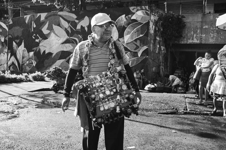 Street vendor selling watches, wearing a cap and striped shirt, with a display board covered in various watches. Background features people and a mural.