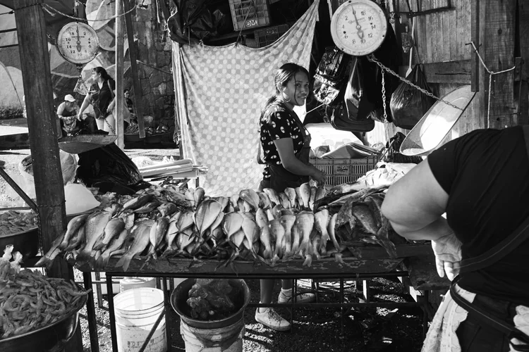Black and white photo of a woman at a seafood market stall with fishes on display on a table and hanging scales.