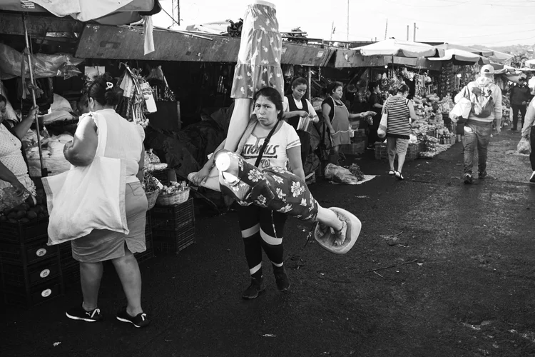 Black and white photo of a busy outdoor market with people shopping and carrying goods; a woman in the foreground holds a mannequin with a dress.