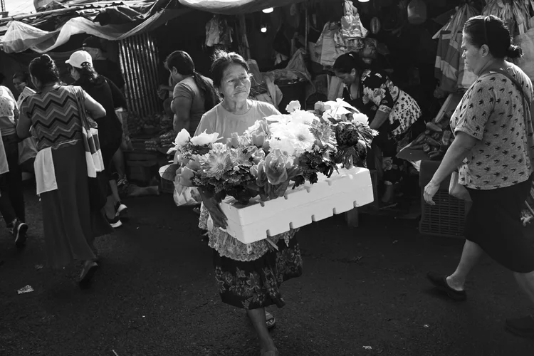 Black and white photo of a woman carrying a box of flowers in a busy market with people walking around.