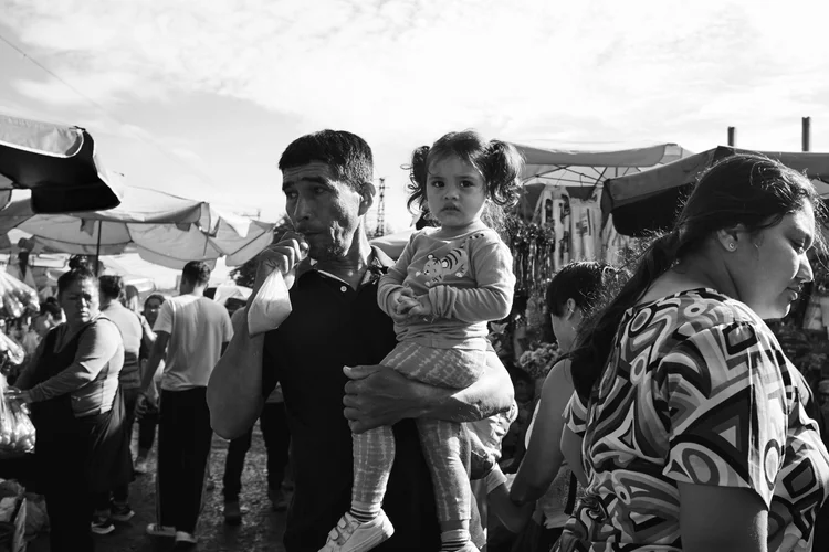 Man holding a child at a crowded outdoor market, surrounded by people and vendor stalls, under a cloudy sky.
