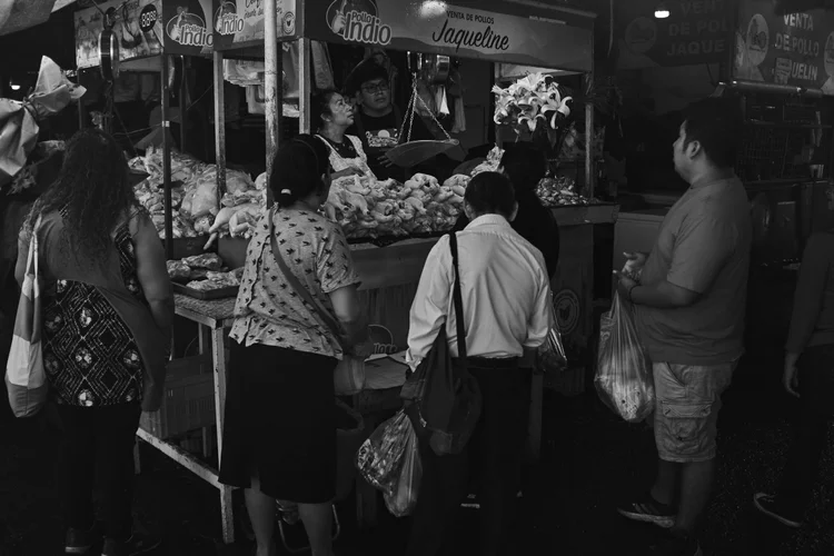Black and white photo of people buying poultry at a market stall with a sign that reads 'Venta de Pollos Jacqueline.' The scene is busy with customers gathered around, and there are flowers visible on the stall.