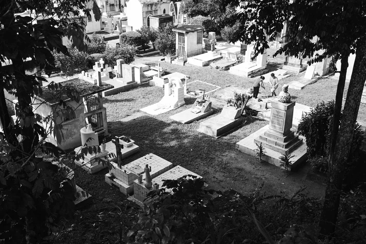 Black and white photo of a cemetery with various tombstones and grave markers, surrounded by trees and shrubs. Two people are walking among the graves.