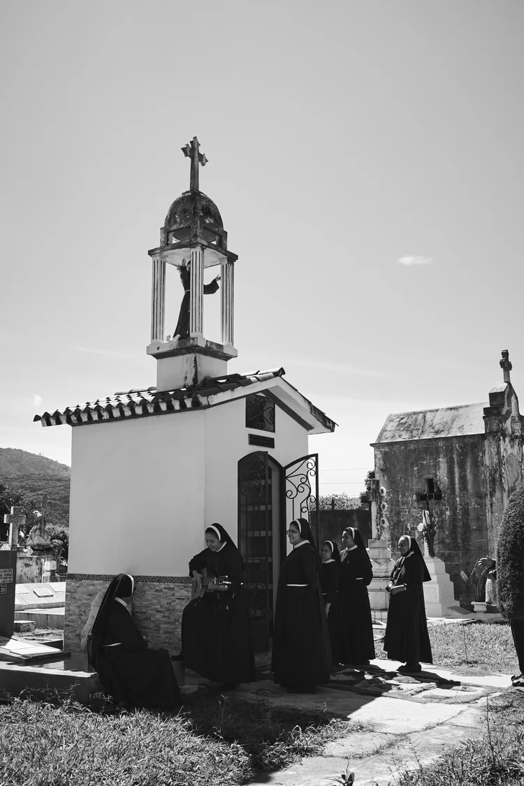 Black and white photo of nuns gathered near a small chapel in a cemetery, with one playing a guitar, surrounded by headstones and crosses.