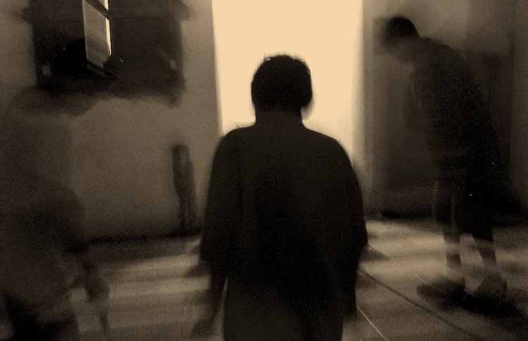 Abstract image of three blurred figures in a dimly lit room with a window.