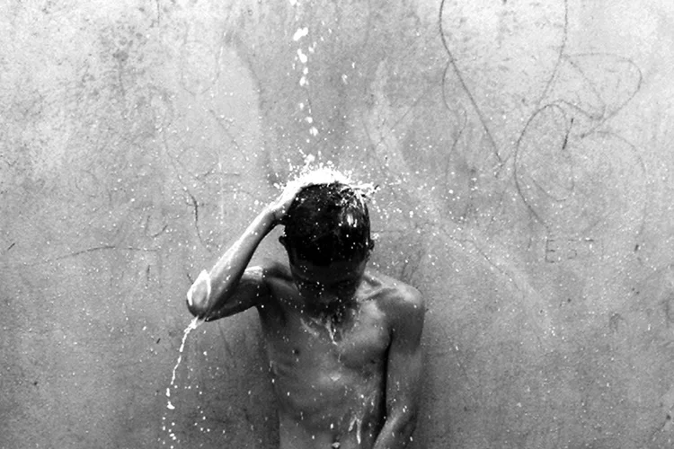 Black and white photo of a person standing under a stream of water against a textured wall.