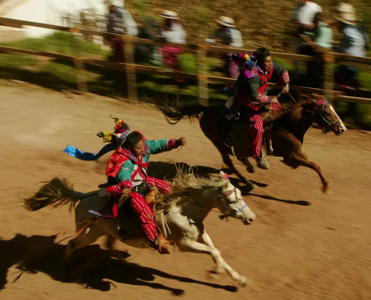 Two people on horseback racing, both wearing traditional colorful clothing and headpieces, on a dirt track with spectators in the background.
