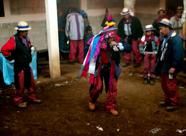 A group of people wearing traditional, colorful clothing with hats gather in a dirt-floored room, participating in a cultural celebration or ritual.