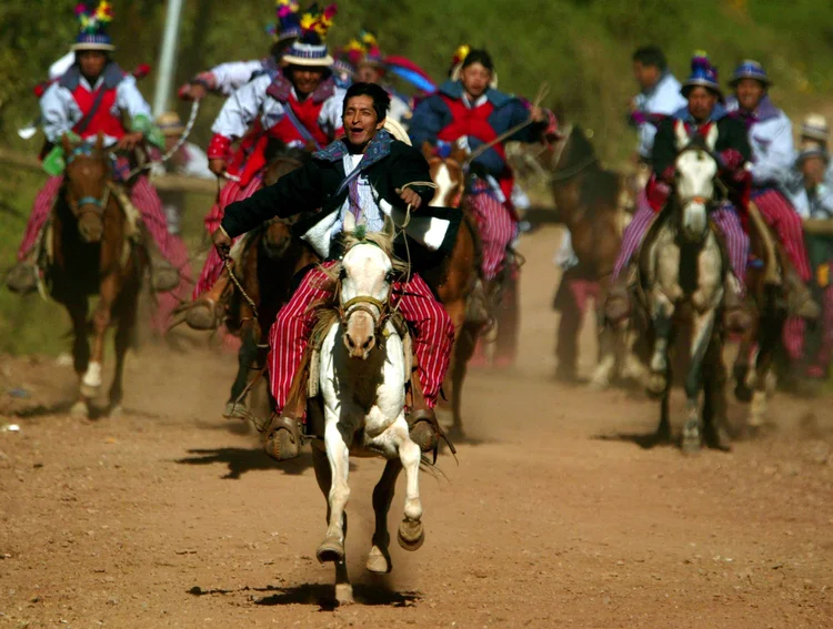 Men in colorful traditional clothing riding horses on a dirt path during a cultural festival or event.