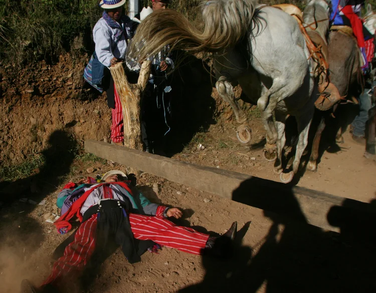 Person in colorful clothing lying on ground with a rearing horse nearby, surrounded by people and rural terrain.
