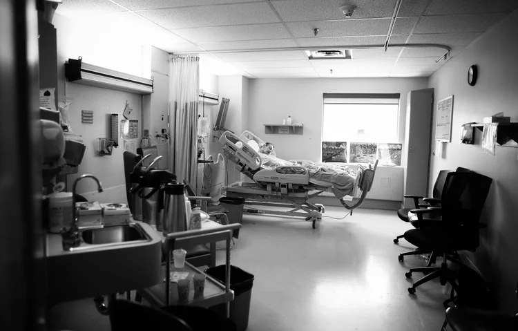 A black and white, wide-angle interior view of a hospital room shows a patient lying in an adjustable bed, positioned towards the center-right. The patient is covered with blankets and appears to be resting. To the left, a medical sink and various supplies are visible on a counter and a rolling cart. Along the left wall, medical equipment and wall-mounted devices are present. The room features a drop ceiling with fluorescent lighting and a window at the far end, partially obscured by blinds, with three framed pictures on the windowsill. On the right side of the room, three office-style chairs are lined against the wall. The overall scene depicts a functional, clinical environment.